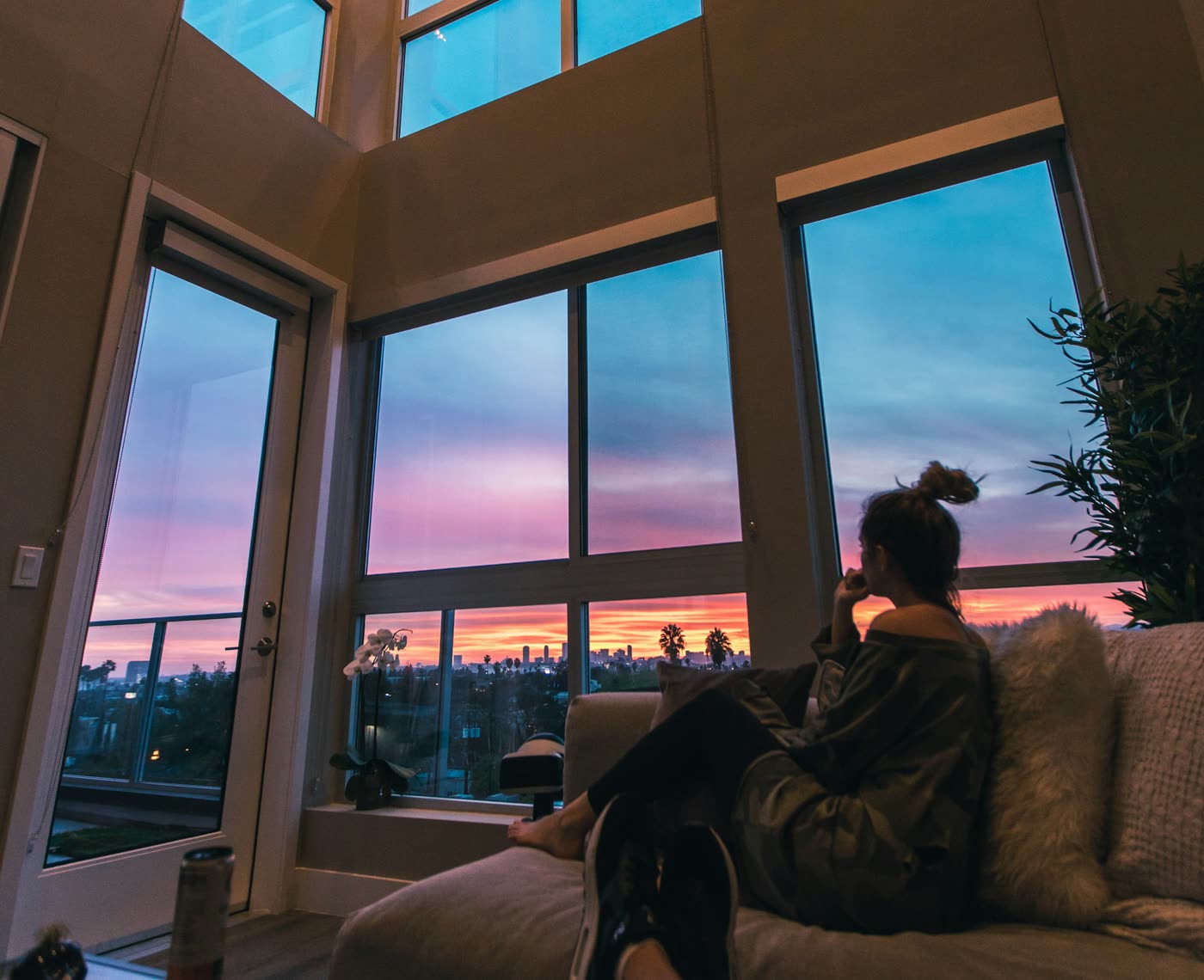 A person relaxing on a sofa by tall windows at sunset